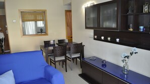 Un salon avec un canapé bleu, une table à manger avec des chaises, un téléviseur fixé au mur et un meuble contenant des objets décoratifs.