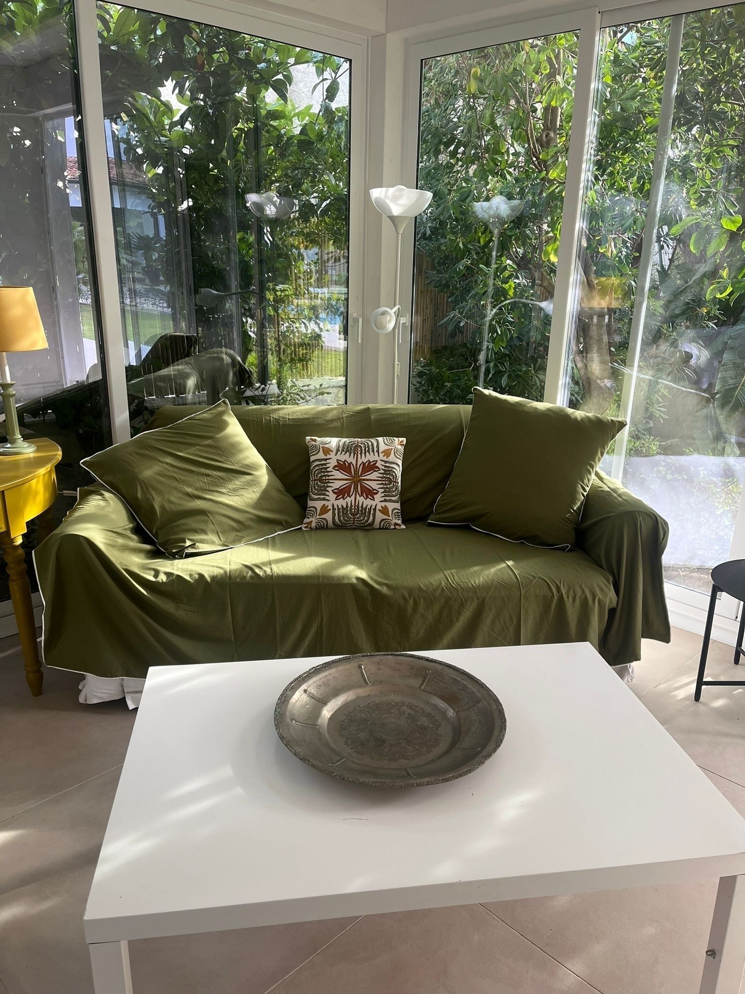 A room with a green sofa, a white table, and a view of greenery through large windows.
