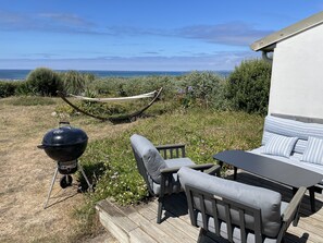 En uteplats med utemöbler, en grill och en hängmatta, med utsikt över en gräsyta och havet.