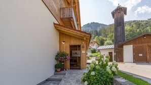 Ein Holzbauten mit einer Steinstufe, die zu einer Tür führt, umgeben von Grünpflanzen vor einer Bergkulisse.