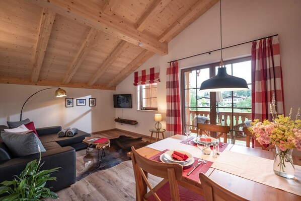 A cozy living room with a wooden ceiling, a sofa, a dining table with chairs, and a view through large windows. - Vacation apartment Wilder Kaiser, 55sqm, balcony (Oberaudorf)