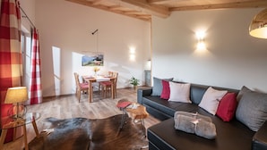 Ein gemütliches Wohnzimmer mit Holzbalkendecke, einer Couch mit Kissen, einem Glastisch und einem Essbereich mit Tisch und Stühlen.