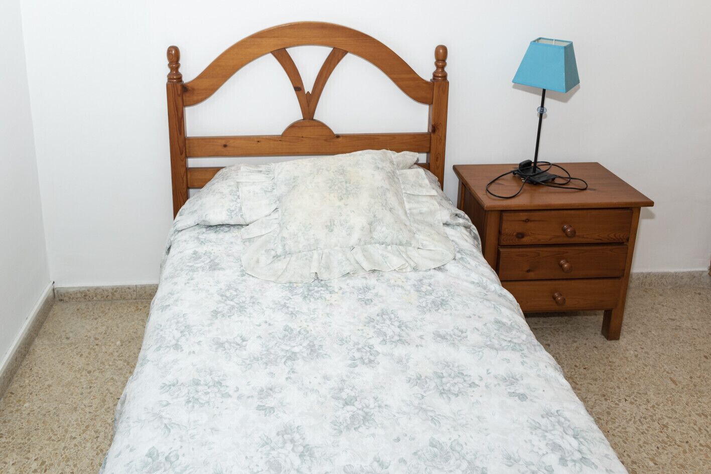 A single bed with a floral bedspread, a wooden headboard, a nightstand with a lamp, and a drawer unit.