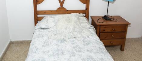 A single bed with a floral bedspread, a wooden headboard, a nightstand with a lamp, and a drawer unit.