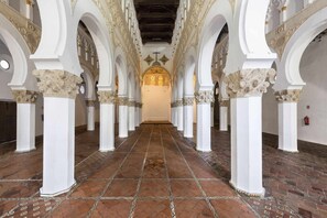 Un pasillo con columnas blancas y suelo estampado, que lleva a una zona iluminada con una cruz.