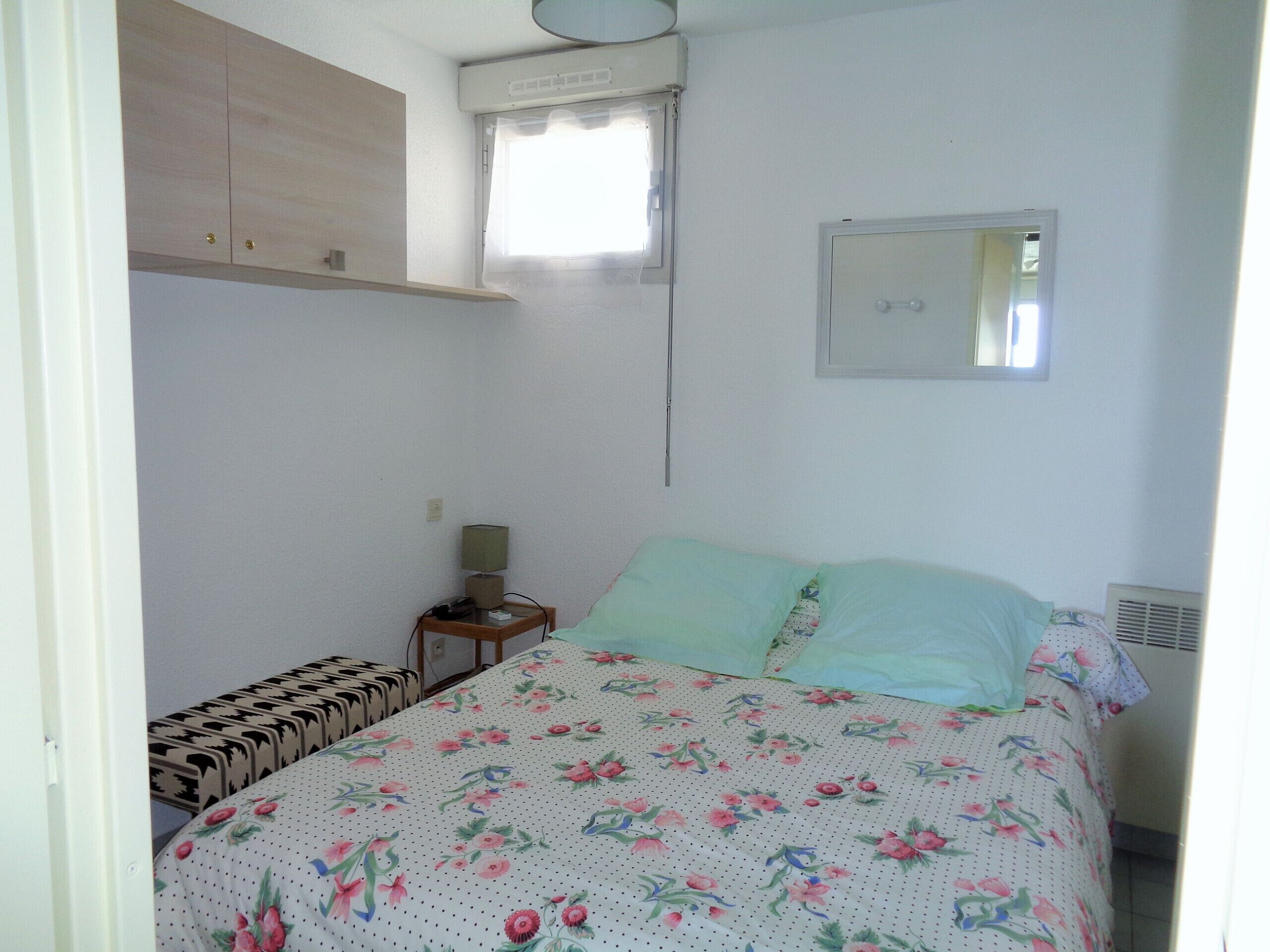 A bedroom with a bed, a bedside table with a lamp, a window with curtains, a mirror, and a wardrobe.