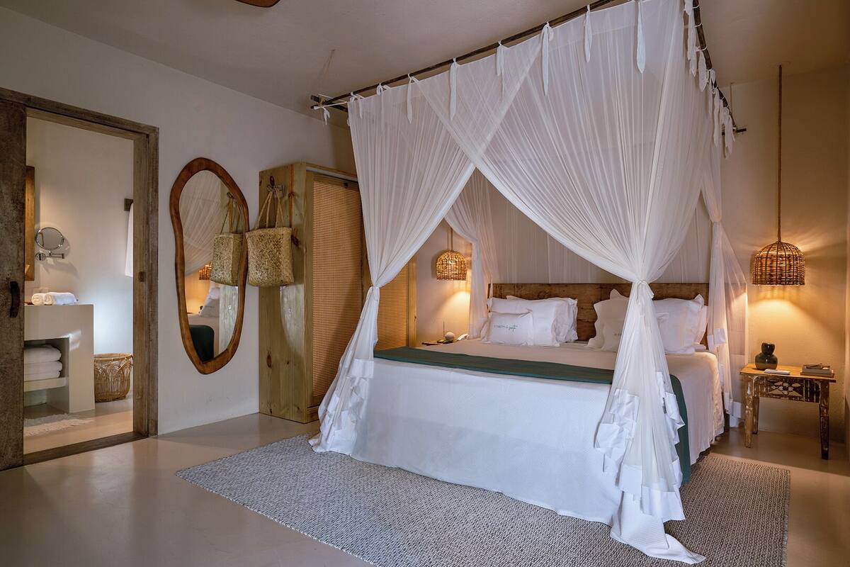 Un dormitorio con cama de dos columnas y dosel, cabecera de madera, mesita de noche con lámpara y un espejo grande en la pared.