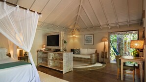 Un dormitorio con cama con dosel, un televisor sobre un mueble de madera, un sofá y vista a la vegetación a través de una ventana.