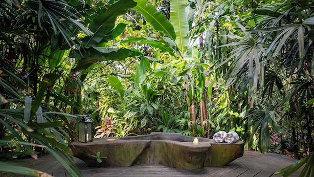 Una terraza de madera rodeada de exuberante vegetación tropical, con un farol y una maceta de piedra con una vela.
