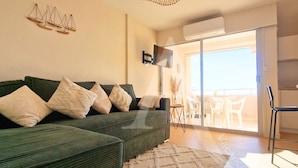 A living room with a green sofa, a flat-screen TV, and a balcony with outdoor furniture.