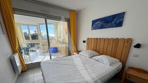 A bedroom with a bed, a wooden headboard, a nightstand, a window with yellow curtains, and a view of the outside balcony.