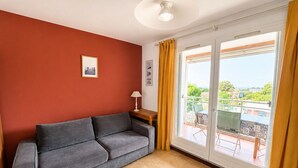 A room with a grey sofa, a small table with a lamp, a framed picture on the wall, and a sliding glass door leading to an outdoor area.
