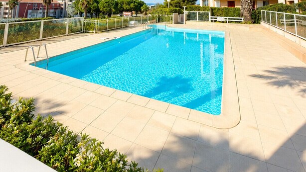 A swimming pool with a clear blue water, surrounded by a tiled deck, palm trees, and a multi-story building with balconies.