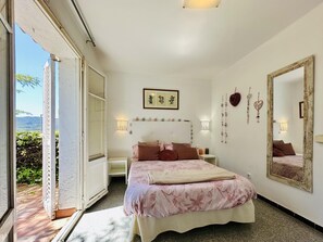 A bedroom with a bed, bedside tables, a large mirror, wall art, and a view through an open door.