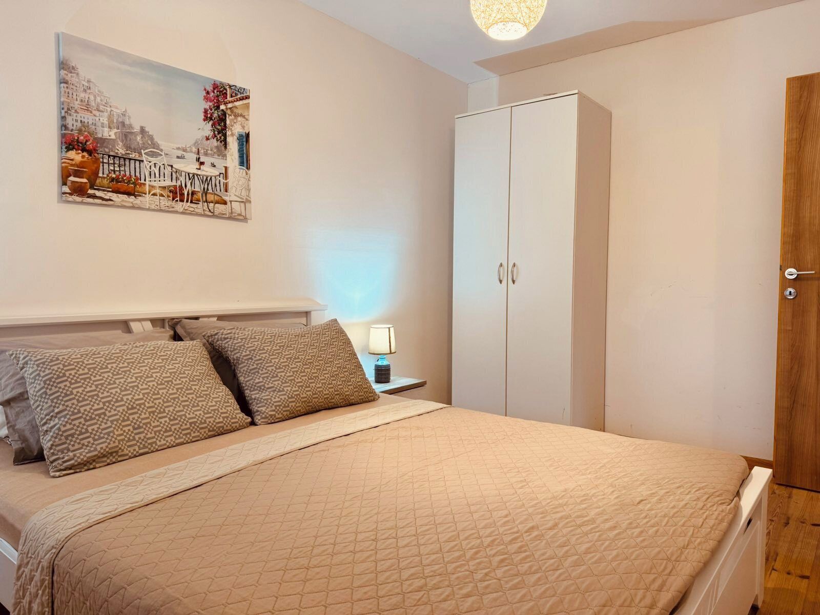 A bedroom with a bed, patterned pillows, a nightstand with a lamp, a wardrobe, and a painting on the wall.