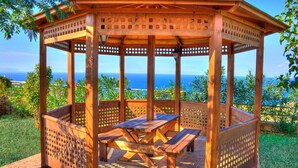 A wooden gazebo with a table and benches, surrounded by a garden and overlooking a scenic view.