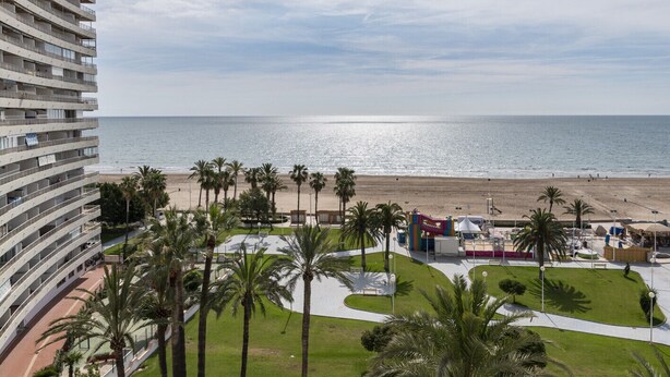 Een hotel aan het strand met meerdere balkons met uitzicht op zee, een zandstrand en palmbomen.