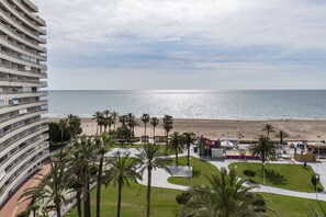 A beachfront hotel with multiple balconies overlooking the sea, a sandy beach, and palm trees.
