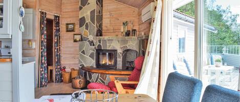 A cozy cabin interior with a stone fireplace, wooden walls, a dining area with a table and chairs, and a view of the outdoors through large windows.