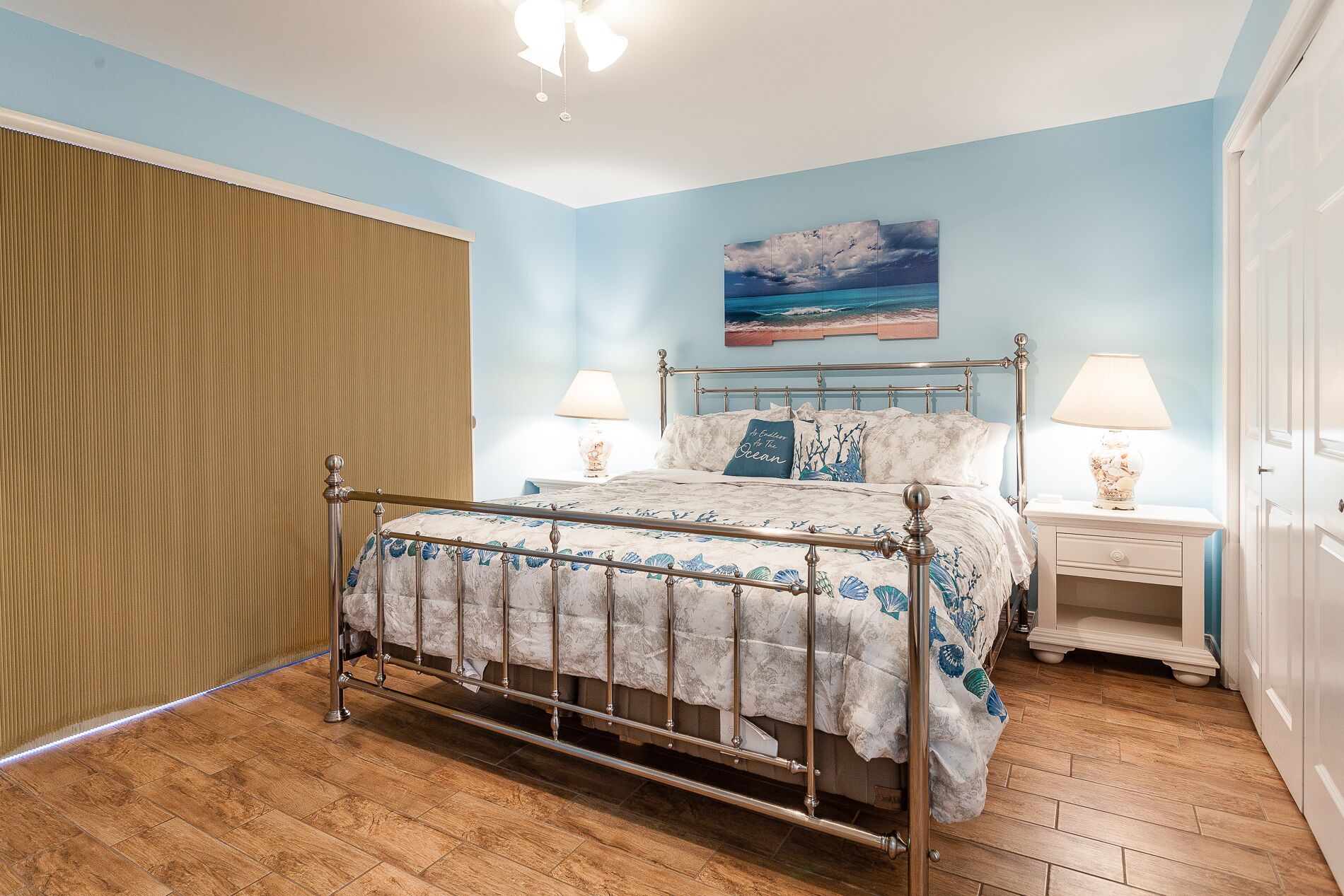 A bedroom with a bed, two bedside tables, a nightstand, a wooden floor, and a window with blinds.