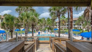 A wooden boardwalk leading to a pool area with palm trees and blue umbrellas. - Highly rated Condo in Ocean Front Community, Wheelchair accessible, Dog friendly (St Augustine Beach)