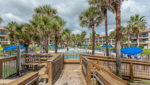 A wooden boardwalk leading to a pool area with palm trees and blue umbrellas. - Highly rated Condo in Ocean Front Community, Wheelchair accessible, Dog friendly (St Augustine Beach)