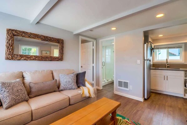 Una sala de estar con un sofá color beige, un banco de madera, un espejo tejido, y una cocina con armarios blancos y un refrigerador de acero inoxidable.