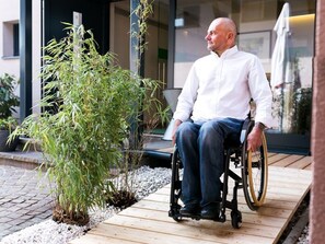 Une personne en fauteuil roulant se trouve sur une terrasse en bois, avec une petite plante à côté d’elle.