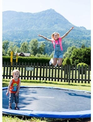 A trampoline with a child jumping and another standing, surrounded by a wooden fence and greenery. - Wheelchair-accessible garden suite with terrace - Landgasthof Hotel Aichingerwirt *** superior (Keuschen)