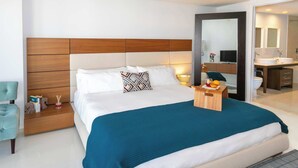 Una habitación de hotel moderna con una cama grande, un cabecero de madera, una mesita de noche con libros y un baño visible a través de una puerta abierta.