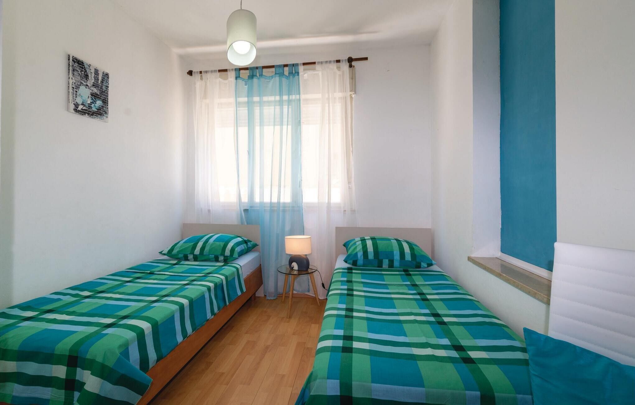 A room with two single beds, a small table with a lamp, and a window with sheer curtains.