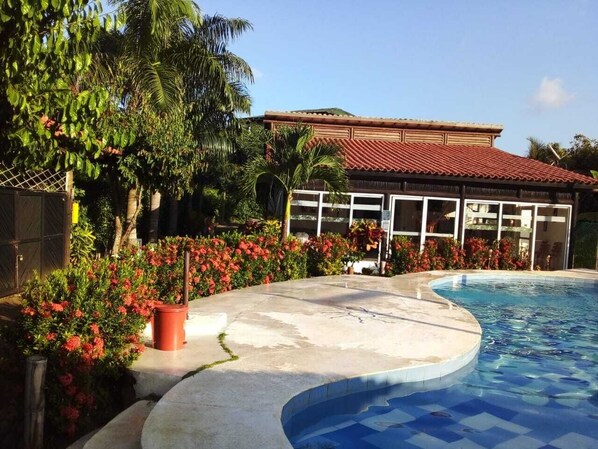A swimming pool with a curved edge, surrounded by a concrete deck. There is a building with a red-tiled roof and large windows in the background.