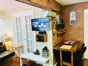 A cozy living room with a TV mounted on a brick wall, a dining table with chairs, and a bench.