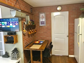 A kitchen with a brick wall, a white refrigerator, a wooden dining table with chairs, a TV mounted on the wall, and a clock above the door. - Nice Apartment/East Hollywood/2 miles to beach/Sleep 6/Free Parking/BBQ/J-7 (Hollywood)