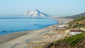 Ein Küstenstrand mit einem Berg im Hintergrund, ein sandiger Uferstreifen und einige Gebäude und Strukturen entlang des Strandes.