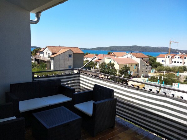 A balcony with wicker furniture, a view of a coastal town, and a clear blue sky.