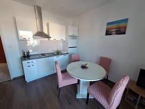 Una cucina moderna con mobili bianchi, un fornello a gas e un bollitore in acciaio inossidabile. È presente un tavolo da pranzo rotondo con sedie rosa e un quadro raffigurante un tramonto sulla parete.