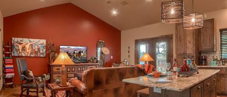 A spacious living room with a red accent wall, a large sofa, a coffee table, and a TV. There is a kitchen area with wooden cabinets and a dining table.