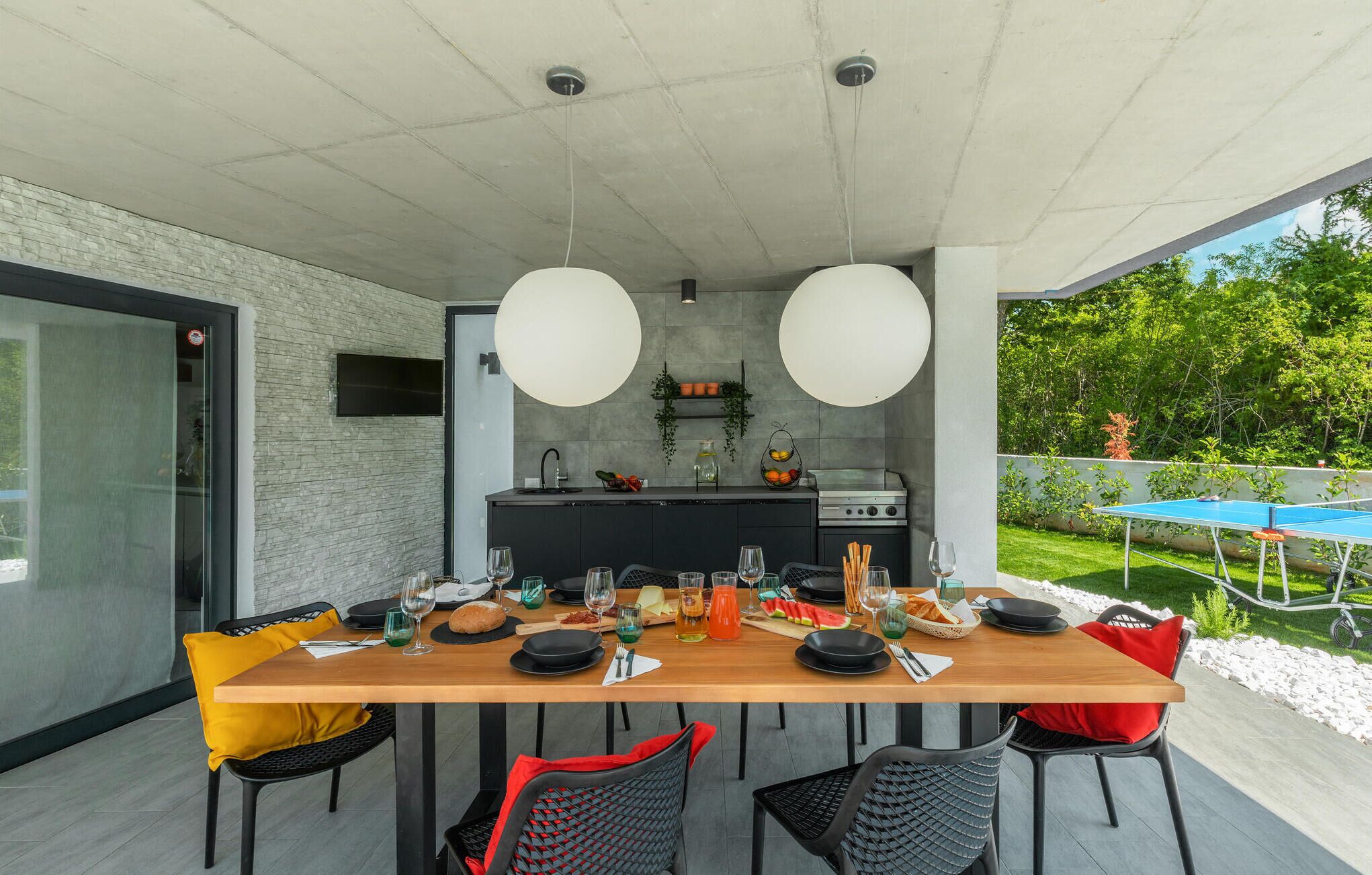 Een moderne eethoek buiten, met een lange houten tafel, zwarte en gele stoelen, en een keuken op de achtergrond.
