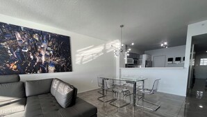 A modern living room with a black leather sofa, a glass dining table with chairs, and a cityscape painting on the wall. - Golden Bay 608, (Sunny Isles Beach)