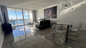 A modern living room with a glass dining table, leather sofa, and a large window overlooking a marina. - Golden Bay 608, (Sunny Isles Beach)