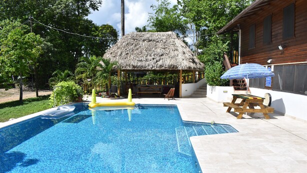 A swimming pool with a thatched-roof structure, a wooden bench, and a table with an umbrella.