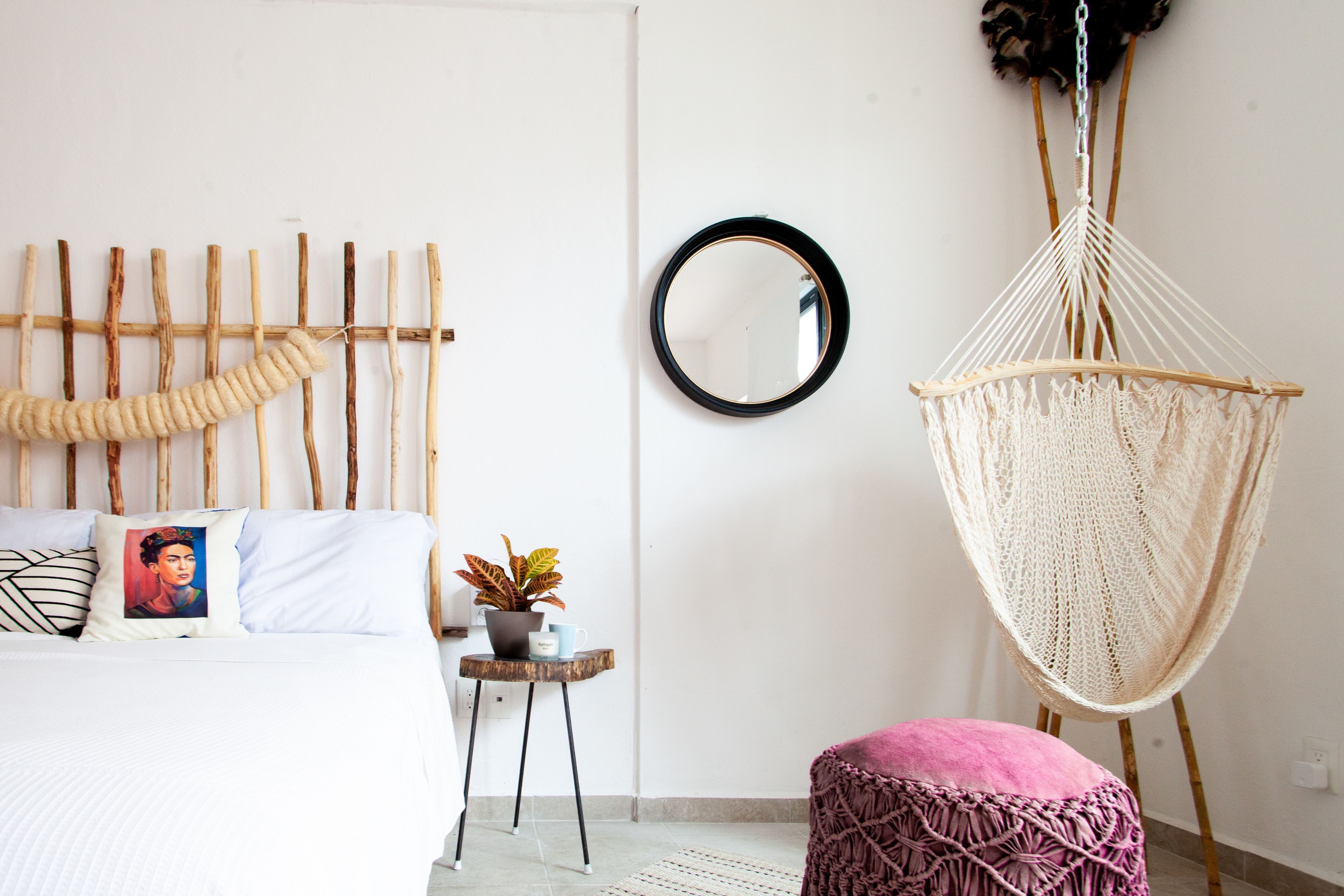 A bedroom with a bed, a bedside table with a plant, a hanging macrame chair, and a round mirror on the wall.