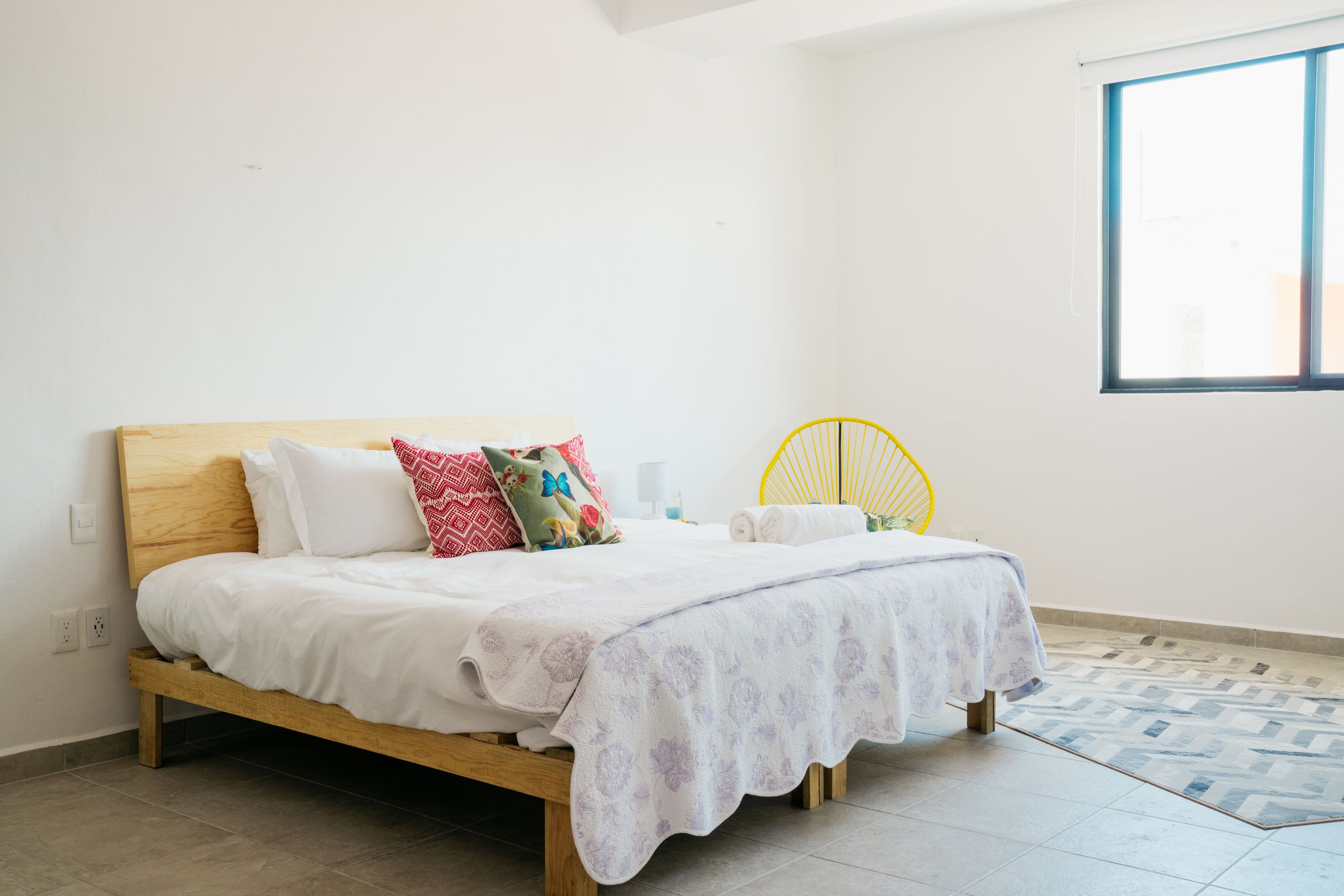 Una cama individual con estructura de madera, ropa de cama blanca y almohadas coloridas. Un sillón amarillo está apoyado contra la pared.