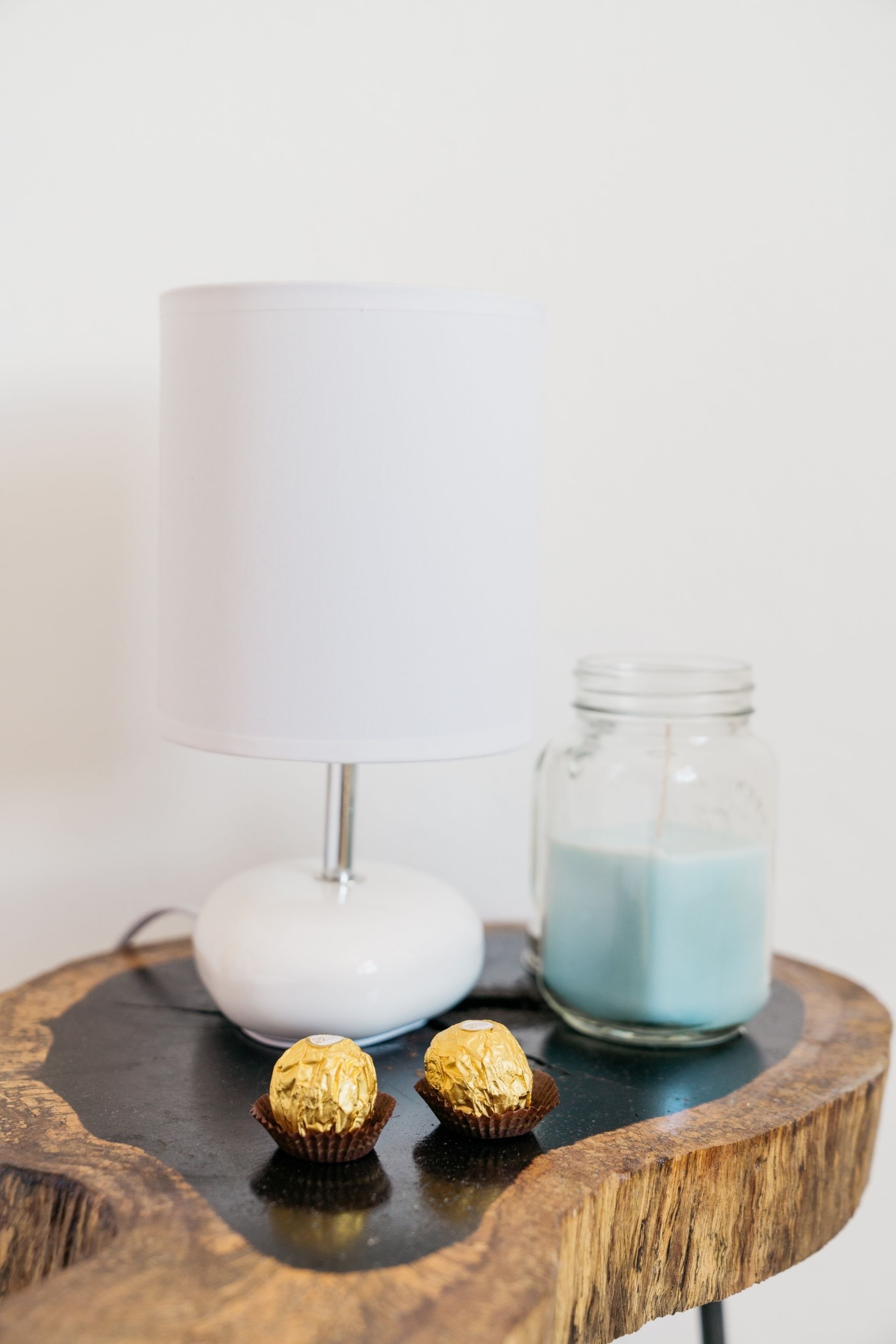A white table lamp, two wrapped chocolates, and a jar of blue candle wax on a round wooden table.