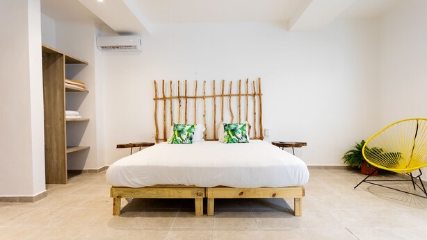 A bedroom with a wooden bed frame, two pillows with tropical prints, a yellow chair, and a small potted plant. - Great Villas Studios & Residences - The Kahlo (Playa del Carmen)