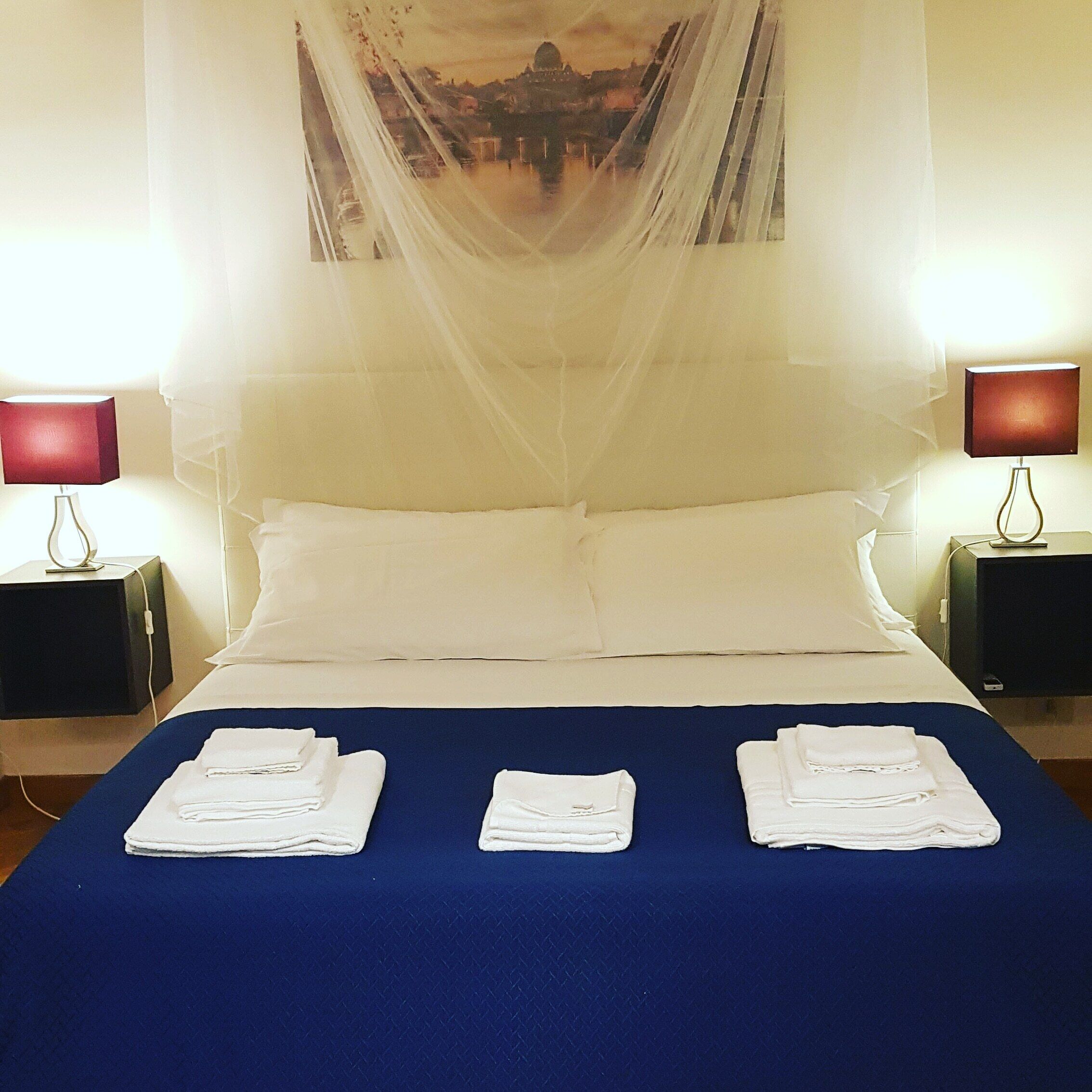 A neatly made bed with white linens and a blue bedspread, two bedside lamps, and a wall-mounted picture.