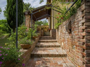 Una scala in mattoni conduce a un'area esterna coperta con una pergola, circondata da rigogliosa vegetazione e piante in vaso.