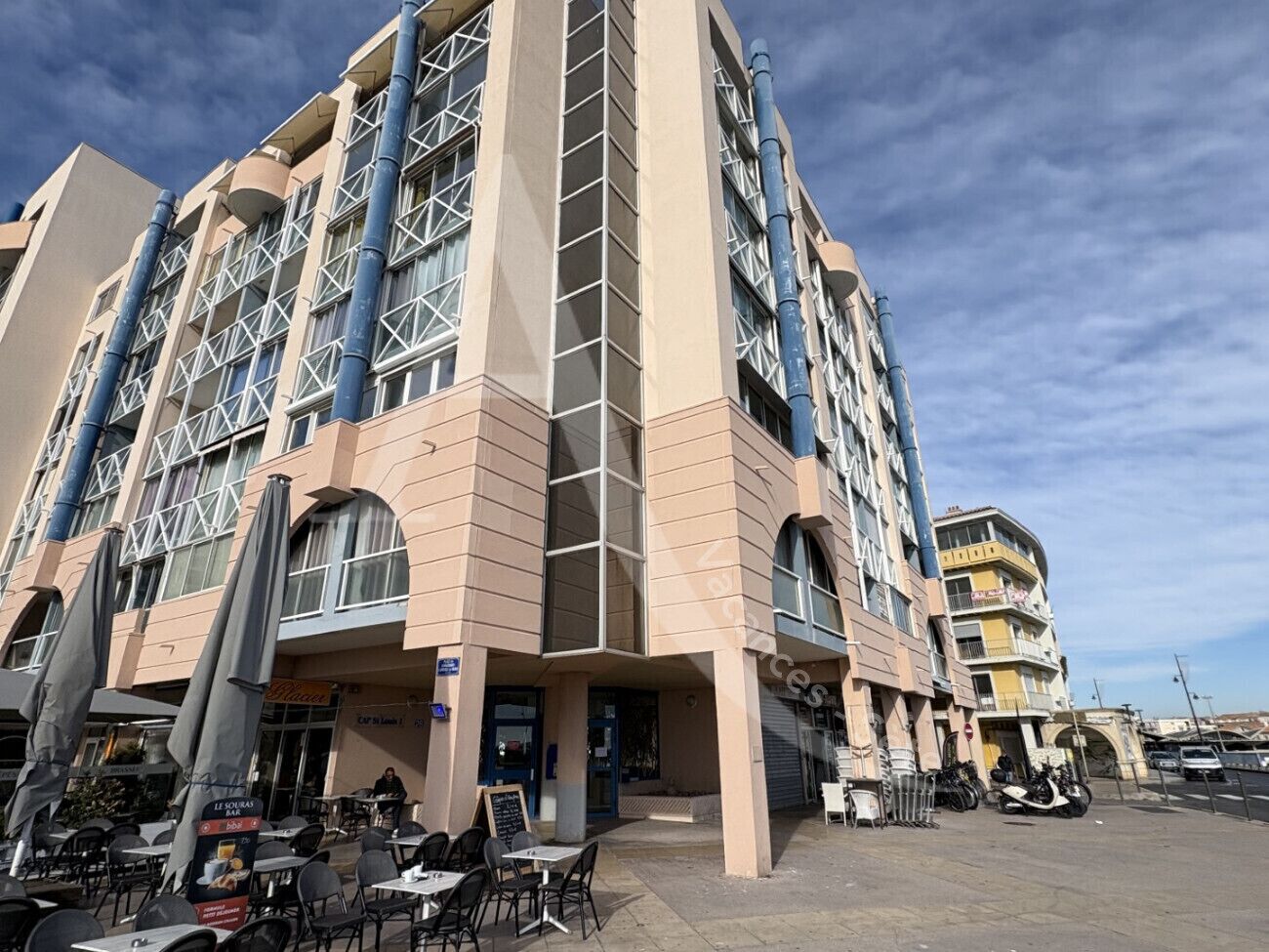 A multi-story hotel with a beige facade, large windows, and a ground-level bar area with outdoor seating.