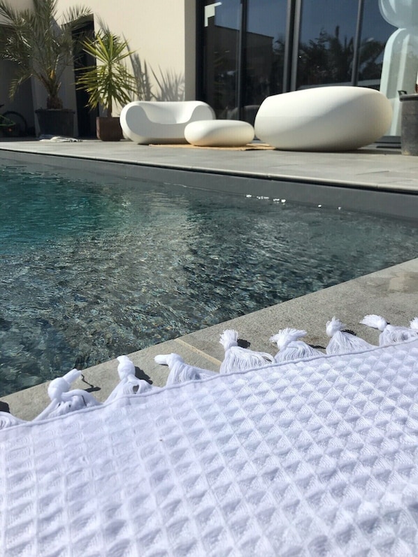 Un área de piscina con agua cristalina, sillas de descanso blancas y una sombrilla texturizada blanca.
